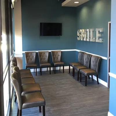 Modern dental waiting room with blue walls, wooden chairs, "SMILE" sign, and wall-mounted TV.