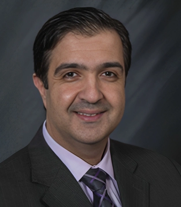 Professional headshot of male dental professional in dark suit and purple tie against gray background.